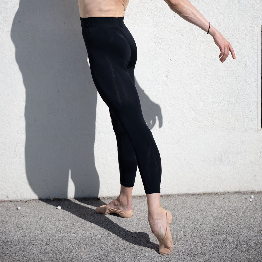 Denys Cherevychko wearing black leggings and beige ballet shoes against a white wall.